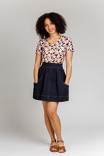Load image into Gallery viewer, Lady stands wearing a denim, short Brumby Skirt with contrasting topstitching detail. Hands in deep scoop pockets.
